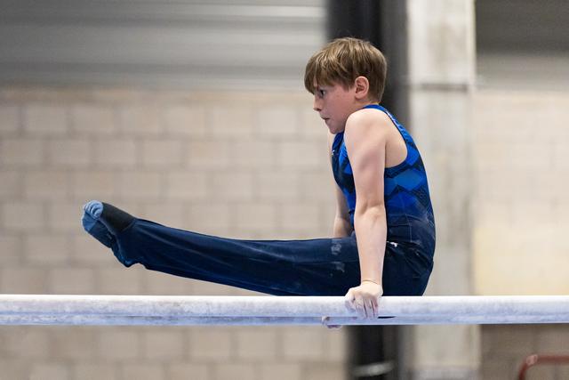 Young gymnast in blue outfit performs on horizontal bar, legs extended parallel to the apparatus in focused concentration