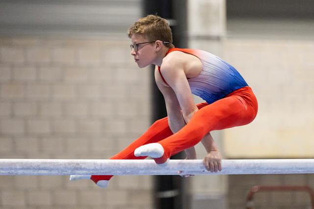 Young male gymnast in orange and blue leotard performs on horizontal bar, body extended horizontally with intense focus