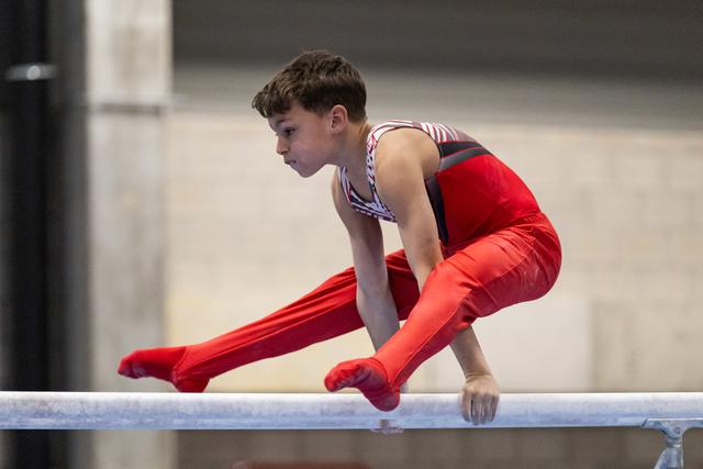 Young gymnast in red uniform performs horizontal bar routine with focused expression and extended leg position