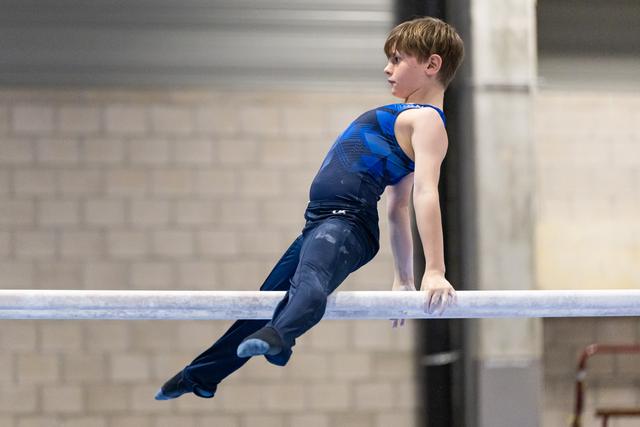 Young male gymnast performing a horizontal bar routine in blue and black competition attire, demonstrating strength and focus