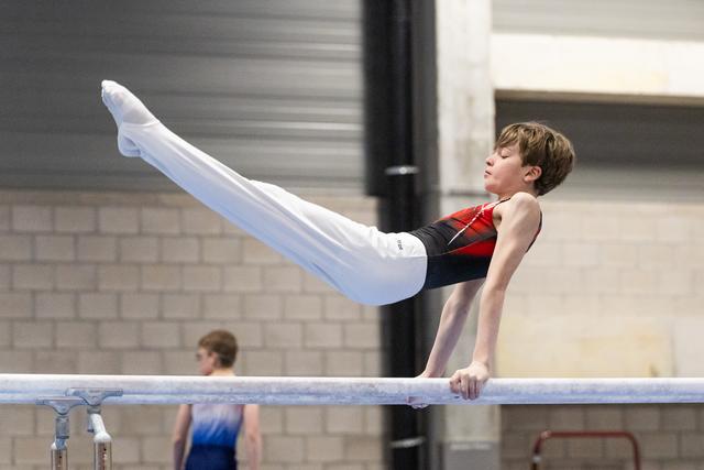 Young male gymnast performs horizontal bar routine with legs extended upward in red and black leotard at indoor gym