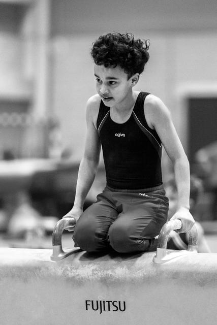 Young gymnast kneeling on balance beam with focused expression, hands gripping apparatus edges during training session