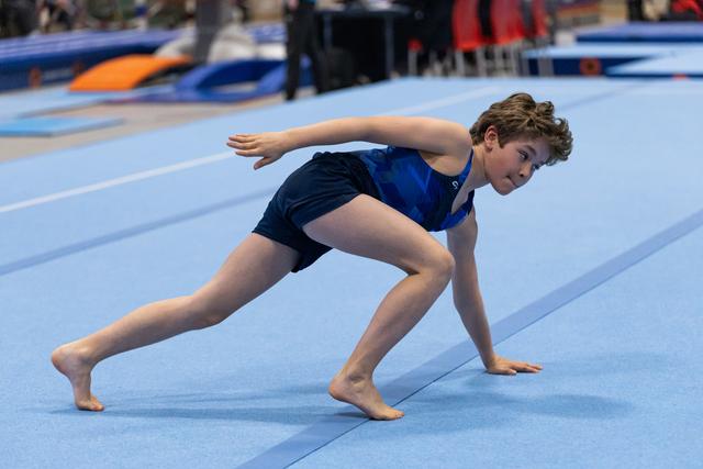 Young gymnast in blue leotard performs a dynamic lunge with extended arms on the spring floor mat