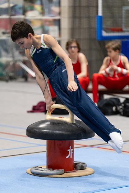 Young gymnast in navy uniform performs horizontal support position on mushroom apparatus while teammates wait in background