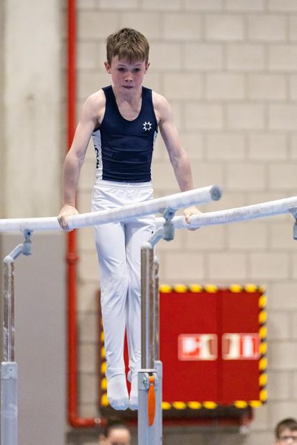 Young male gymnast performs on parallel bars wearing navy and white, displaying intense concentration in indoor gymnasium
