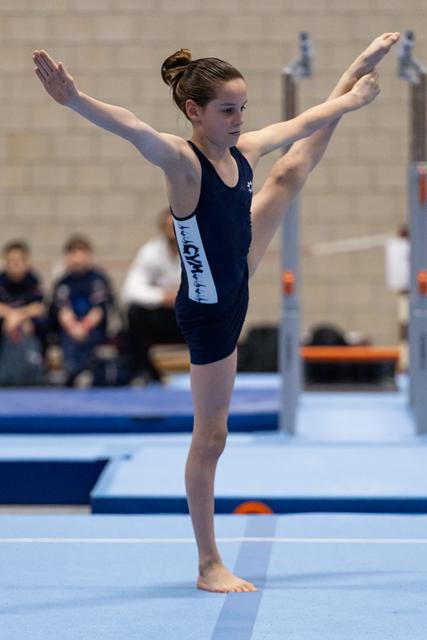 Young gymnast in navy leotard performs an expressive floor routine pose with arms raised, displaying focus and grace