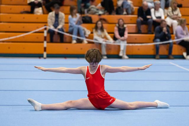 Young gymnast in red leotard performs split on floor mat with arms extended, spectators watching from wooden bleachers