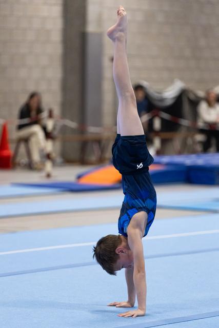 Young gymnast in blue leotard executes controlled handstand with pointed toes on blue floor mat during routine