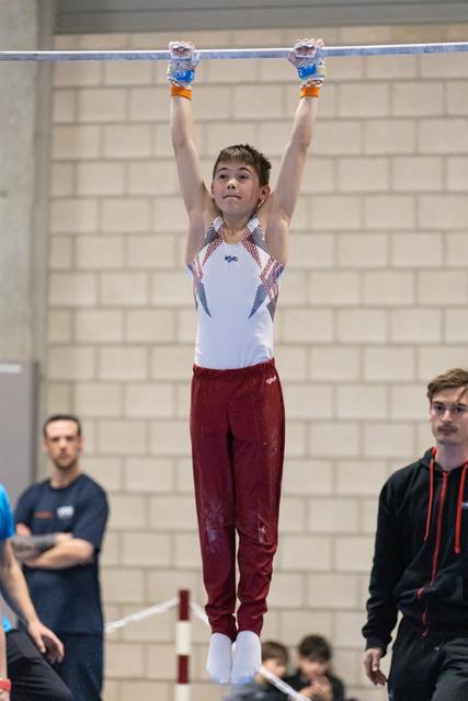 Young male gymnast gripping high bar mid-routine, focused expression, wearing maroon pants and white leotard in indoor gym