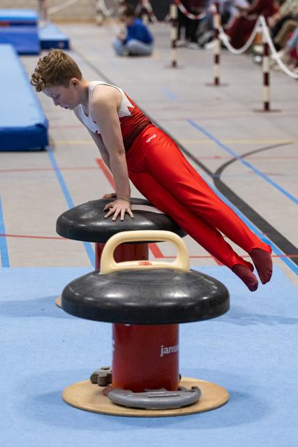 Young gymnast in red uniform executes a strength hold on the pommel horse, body extended horizontally during training.