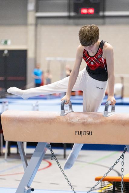 Young male gymnast executing a pommel horse routine with white pants and red-black leotard at indoor gymnastics facility