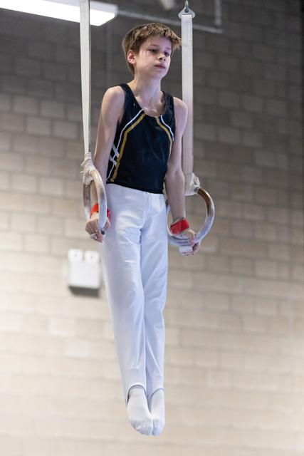 Young male gymnast executing a still rings routine in white pants and navy leotard, displaying focused concentration