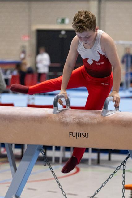 Young gymnast in red and white performs on pommel horse with intense concentration, leg extended during training