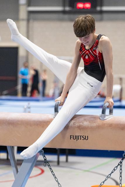 Young male gymnast performing on pommel horse with legs extended upward, wearing red and black leotard in training facility