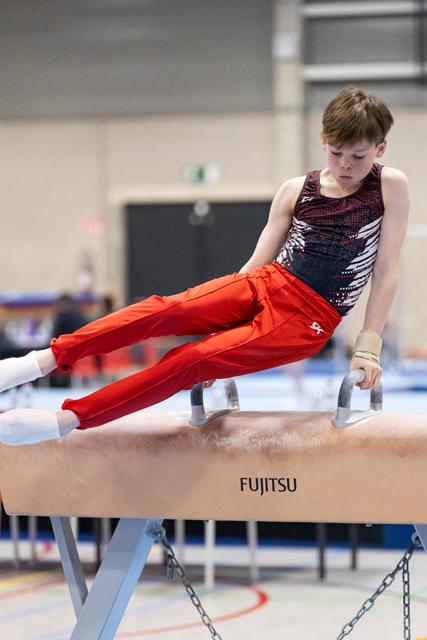 Young male gymnast performs pommel horse routine in red pants and patterned leotard, displaying intense concentration