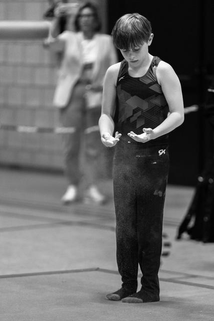 Young gymnast in black leotard concentrates while chalking hands before routine, coach visible in background