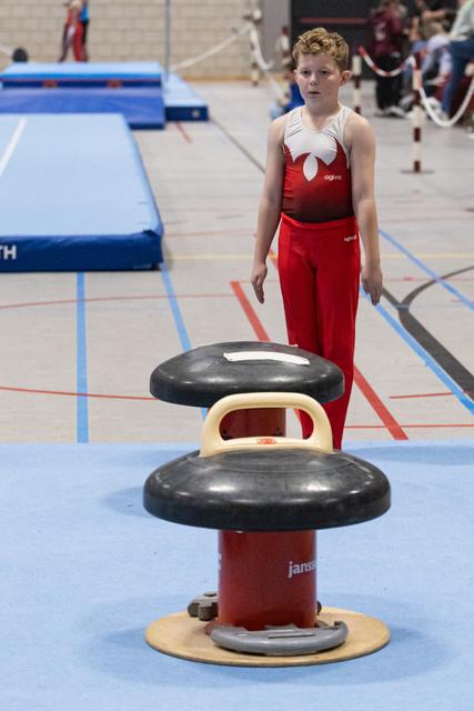 Young gymnast in red uniform stands ready behind a vault apparatus, focused and composed before their turn
