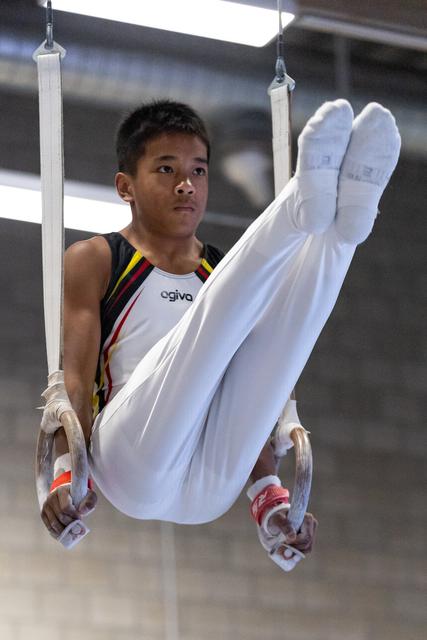 Young gymnast performing an L-sit position on rings, displaying focus and control during his routine at indoor training facility