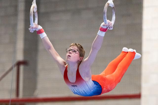 Young gymnast in orange pants and gradient leotard performs rings routine with extended body position, wearing glasses