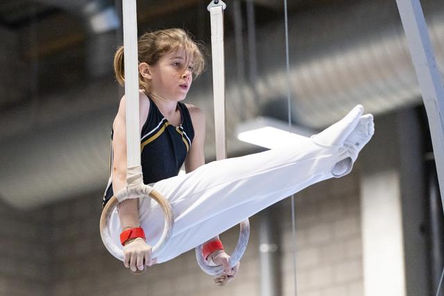 Young gymnast performs on rings wearing white pants and dark leotard, demonstrating focus and determination during apparatus work