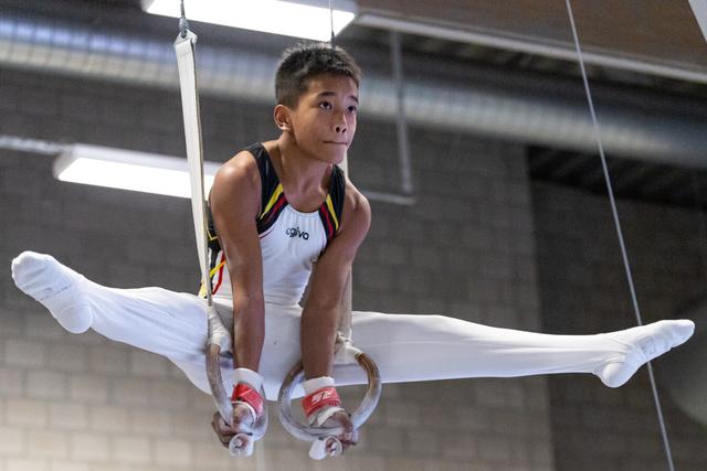 Young male gymnast performing a straddle hold on rings, focused expression, white uniform with colorful trim, indoor gym