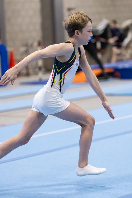 Young gymnast in white and yellow leotard runs across blue mat during floor routine, arms extended in focused form