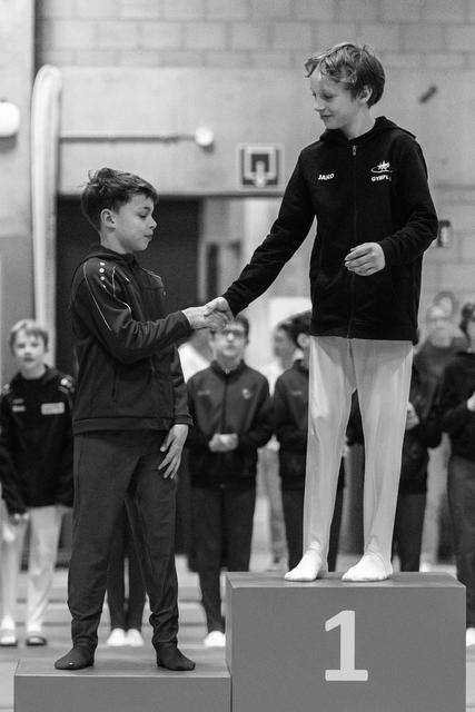 Young gymnast in black shakes hands with first-place winner on podium while teammates watch in gymnasium background
