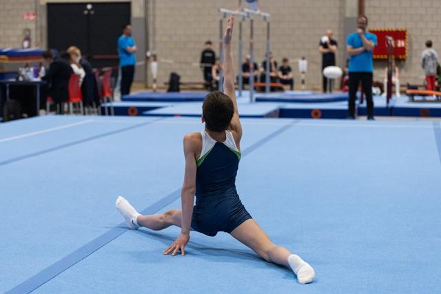 Young gymnast performs splits stretch on blue floor mat during training, arms raised overhead in focused preparation pose