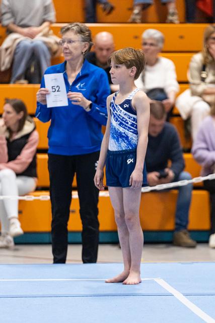 Young gymnast in blue competition attire stands at attention on floor mat while judge holds score card behind him