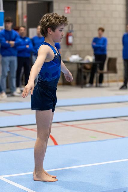 Young gymnast in blue leotard stands barefoot on floor mat, arms positioned, curly hair, focused expression, teammates in background