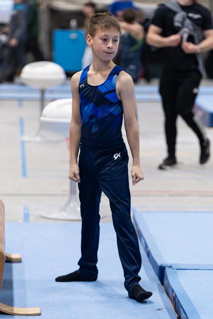 Young male gymnast in blue geometric leotard stands ready on competition floor, focused expression before routine