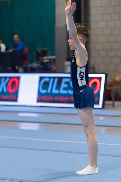 Young gymnast in navy leotard stands at attention with arms raised overhead on blue floor mat before routine