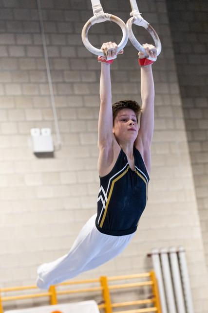 Young gymnast performing a straight-body hold on still rings, arms extended overhead, wearing navy and gold leotard