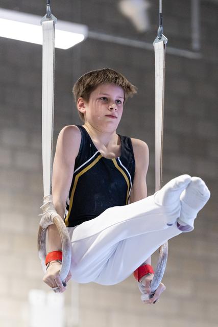 Young gymnast performing L-sit hold on still rings, displaying intense focus and strength in black leotard with gold trim
