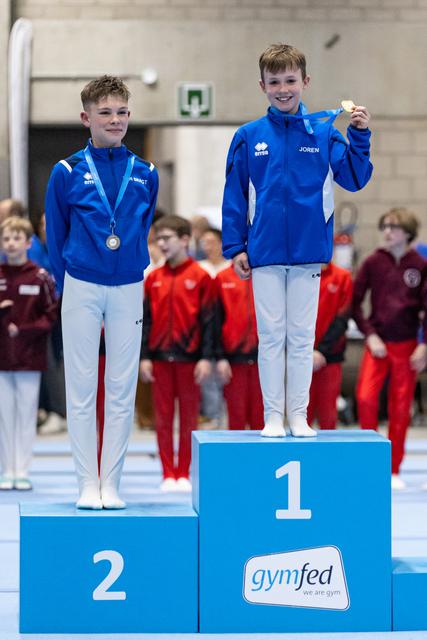 Two young gymnasts in blue tracksuits stand on medal podium, first and second place winners displaying their medals with pride