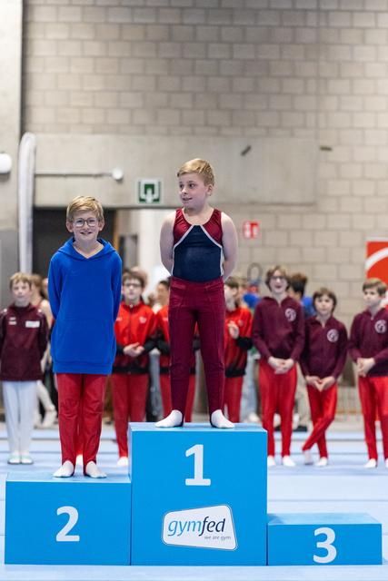 Two young gymnasts stand on a podium in red and blue uniforms, celebrating their achievements with teammates watching behind