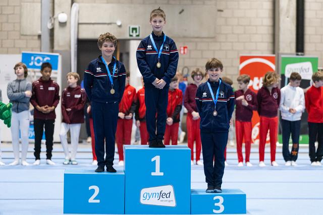 Three young gymnasts stand on victory podium wearing medals and matching navy tracksuits as teammates watch in background