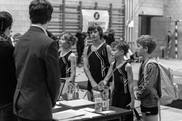 Four young gymnasts in black leotards listen attentively to their coach near a table with water bottles in a gymnasium