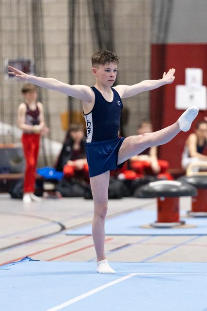 Young gymnast in navy leotard performs an arabesque balance on floor mat, arms extended, focused expression