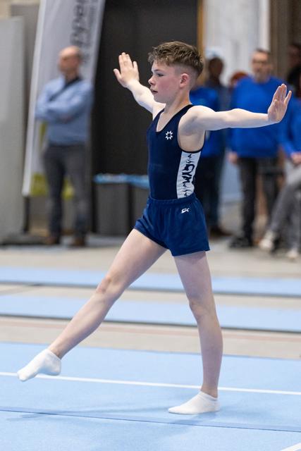 Young gymnast performs floor exercise with extended arms in navy uniform at indoor gymnastics meet