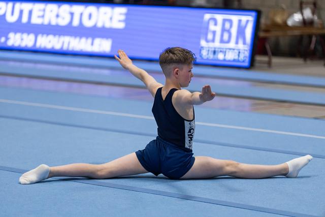 Young male gymnast performs a splits pose on the blue floor mat with arms extended, demonstrating flexibility and form