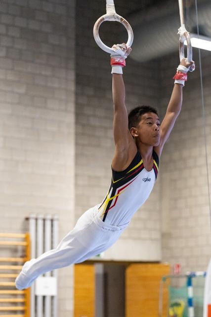 Young gymnast in white and yellow leotard performs a controlled hold on still rings, arms extended above