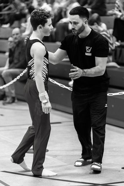 Coach offering encouragement to young gymnast in striped leotard, hand on shoulder in supportive gesture during indoor meet