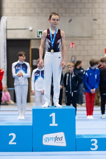 Young gymnast in maroon leotard stands on first-place podium wearing gold medal and blue ribbon at gymnastics meet