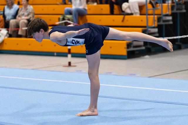 Young gymnast performs a horizontal balance pose on the floor exercise mat with spectators watching from the bleachers