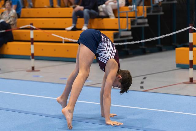 Young gymnast performs a bridge backbend on blue floor mat during gymnastics event with spectators in background