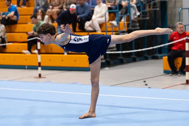 Young gymnast performs an elegant arabesque balance during floor routine at indoor meet with spectators in bleachers