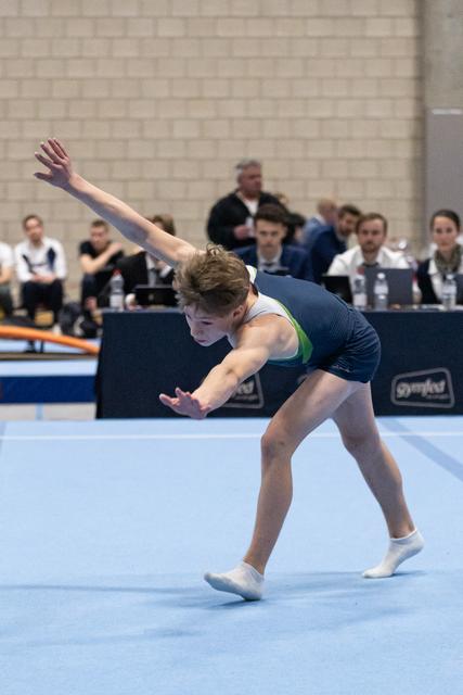 Young male gymnast performs expressive floor routine with extended arm pose during competitive meet