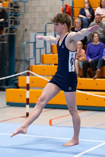 Male gymnast in navy leotard performs floor routine with arms extended, displaying focus and form before spectators