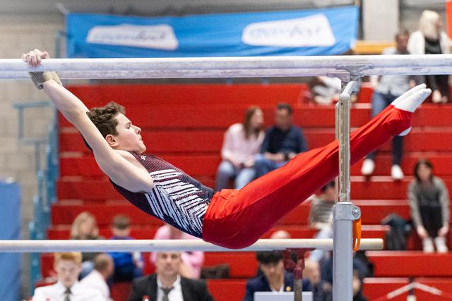 Male gymnast performs horizontal bar routine with extended body position over red bleachers filled with spectators